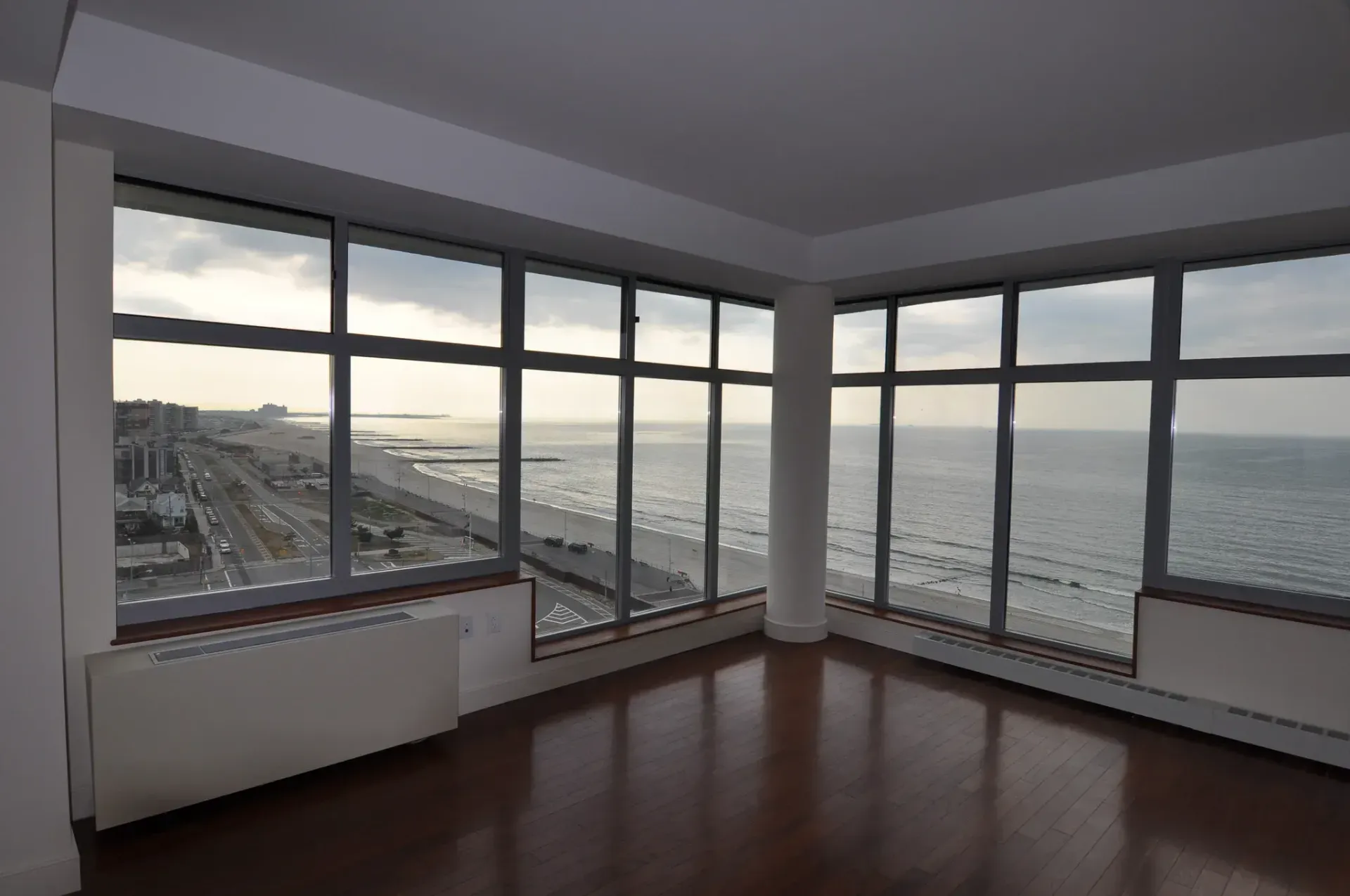 Empty room with large windows overlooking a beach and ocean. Brown wood floors, white walls and a central pillar.