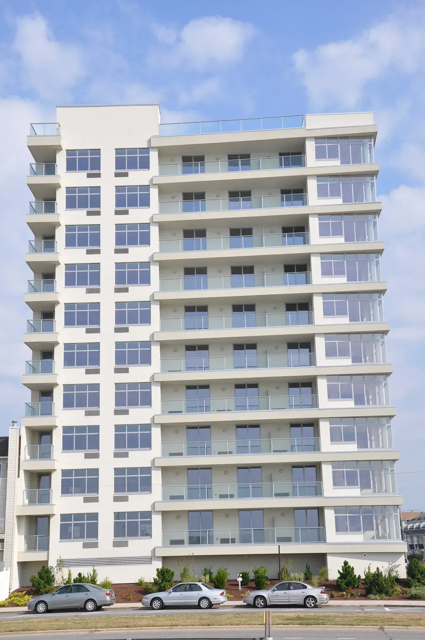 A tall white apartment building with many balconies and windows stands in front of a partly cloudy sky