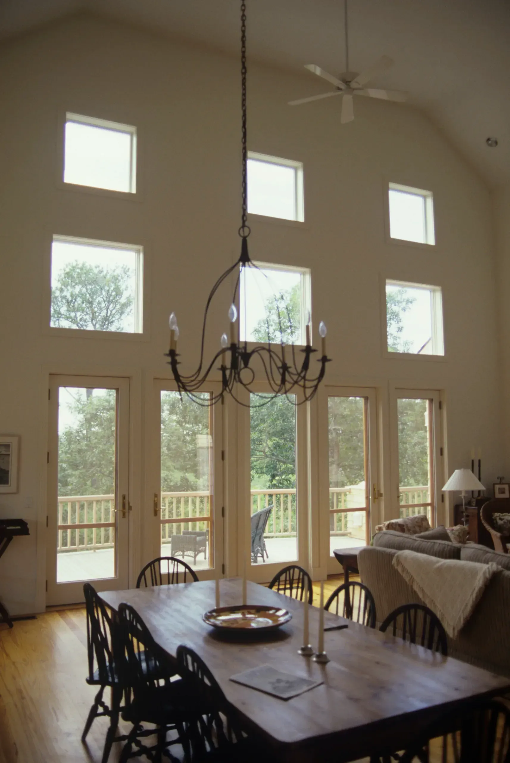 A dining room with a long wooden table, chairs, chandelier, and tall windows overlooking a deck and trees.