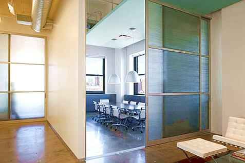 Office space with a glass-walled conference room. Light-colored walls and gray flooring. White furniture.