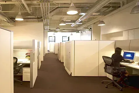 Office cubicles in a long row with a person working at a computer; neutral tones, overhead lighting.