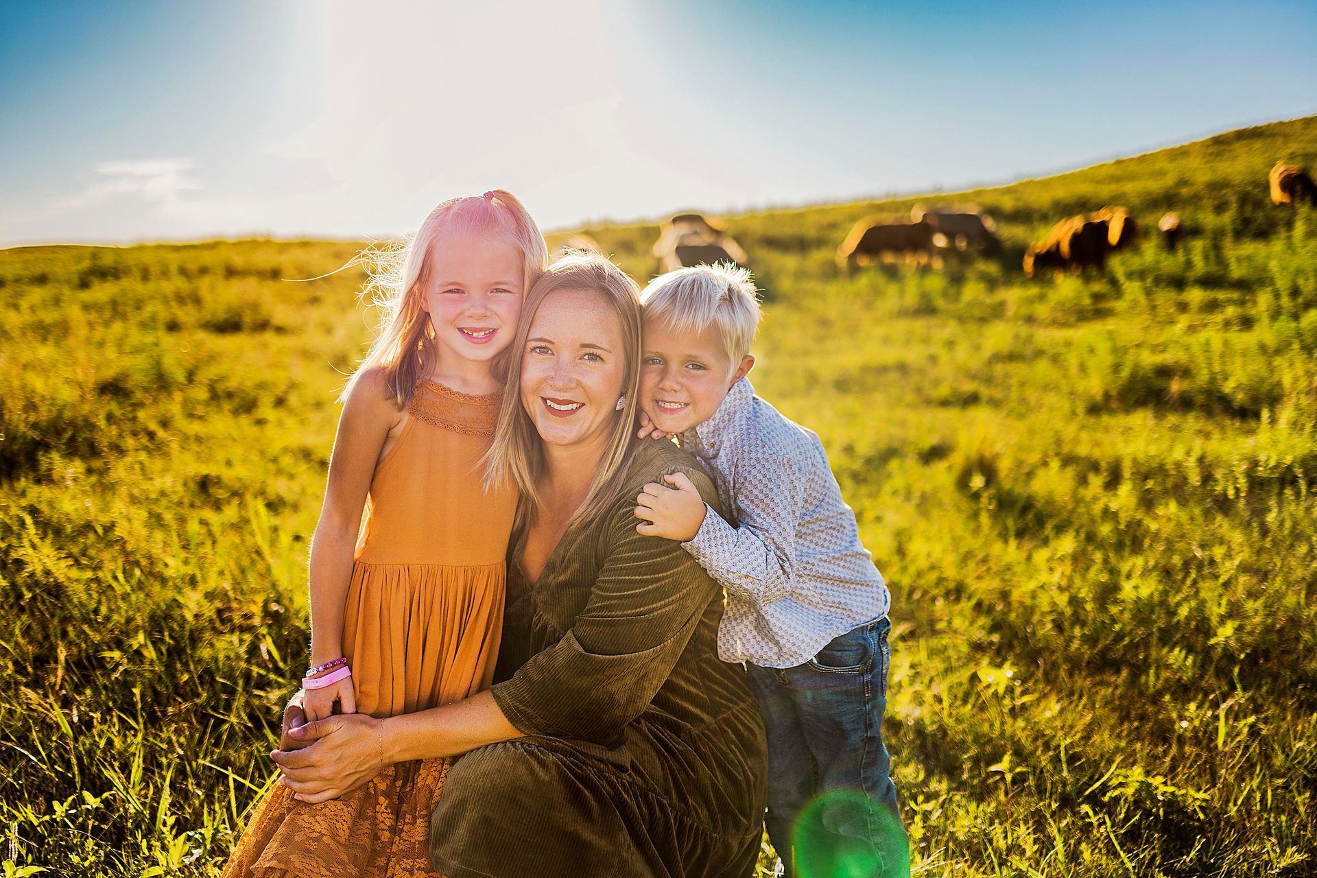 Woman and two children embrace in a sunny field. The woman is smiling, kids are hugging her. Cows graze in the background.