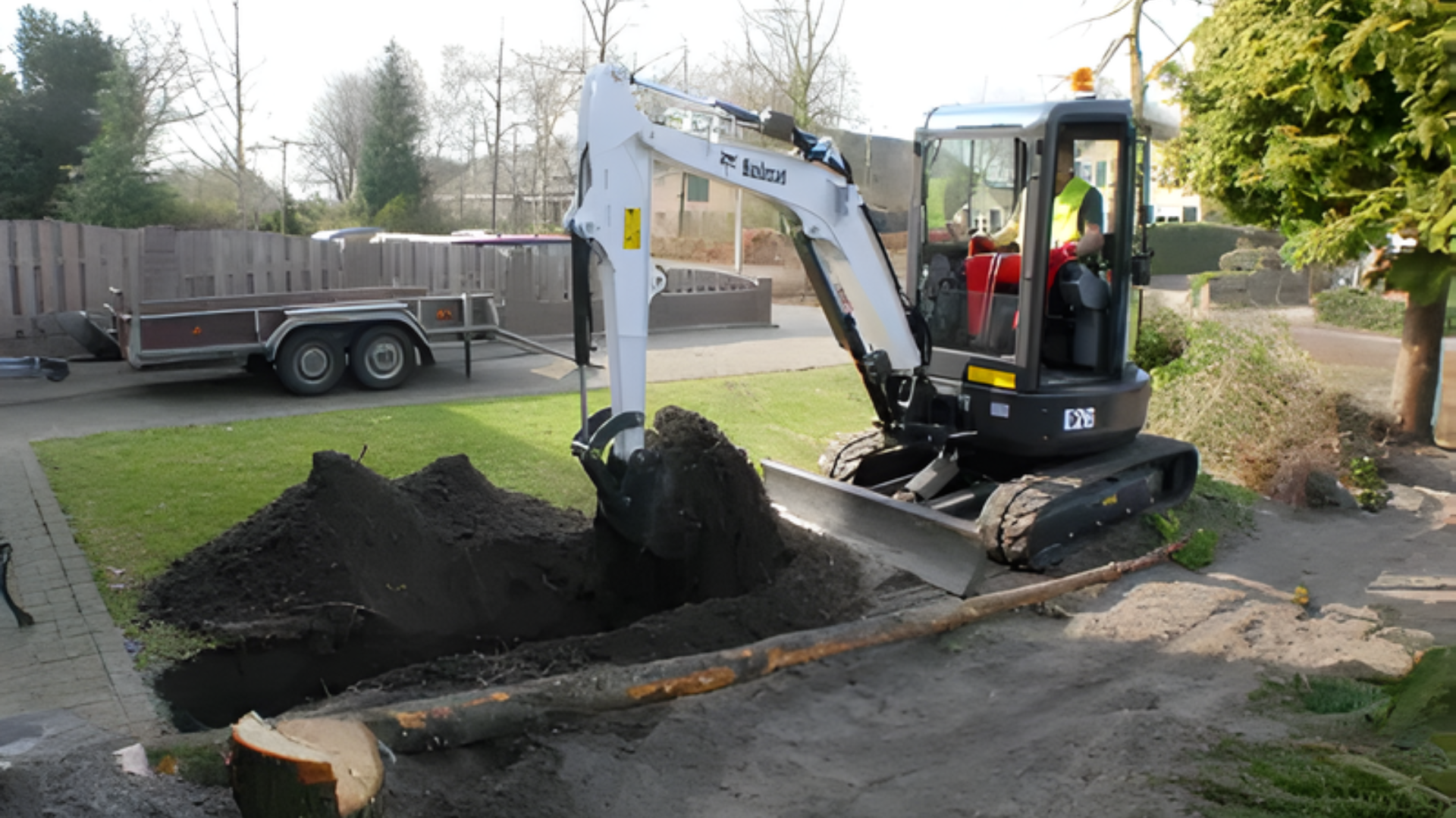 Mini excavator digging in a yard, moving dark soil. A trailer is nearby.