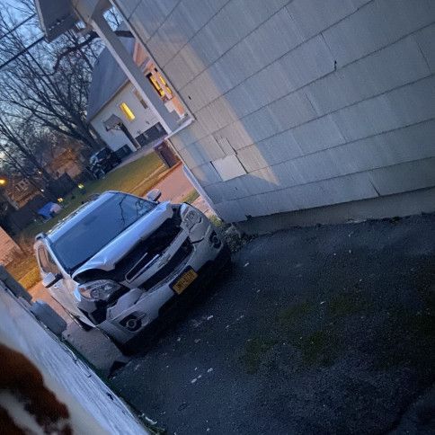 A white car with a damaged front end is parked in front of a house.