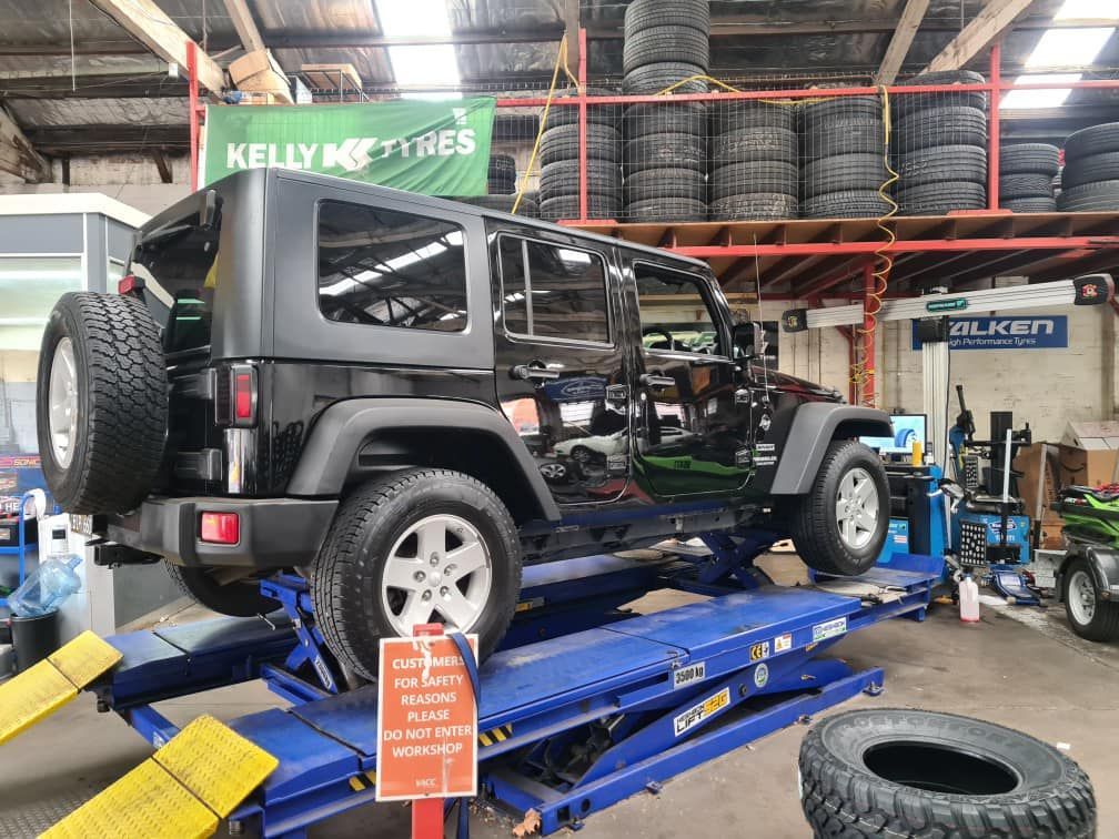 Black Jeep Wrangler on a blue hydraulic lift inside a tire shop. Spare tire mounted.