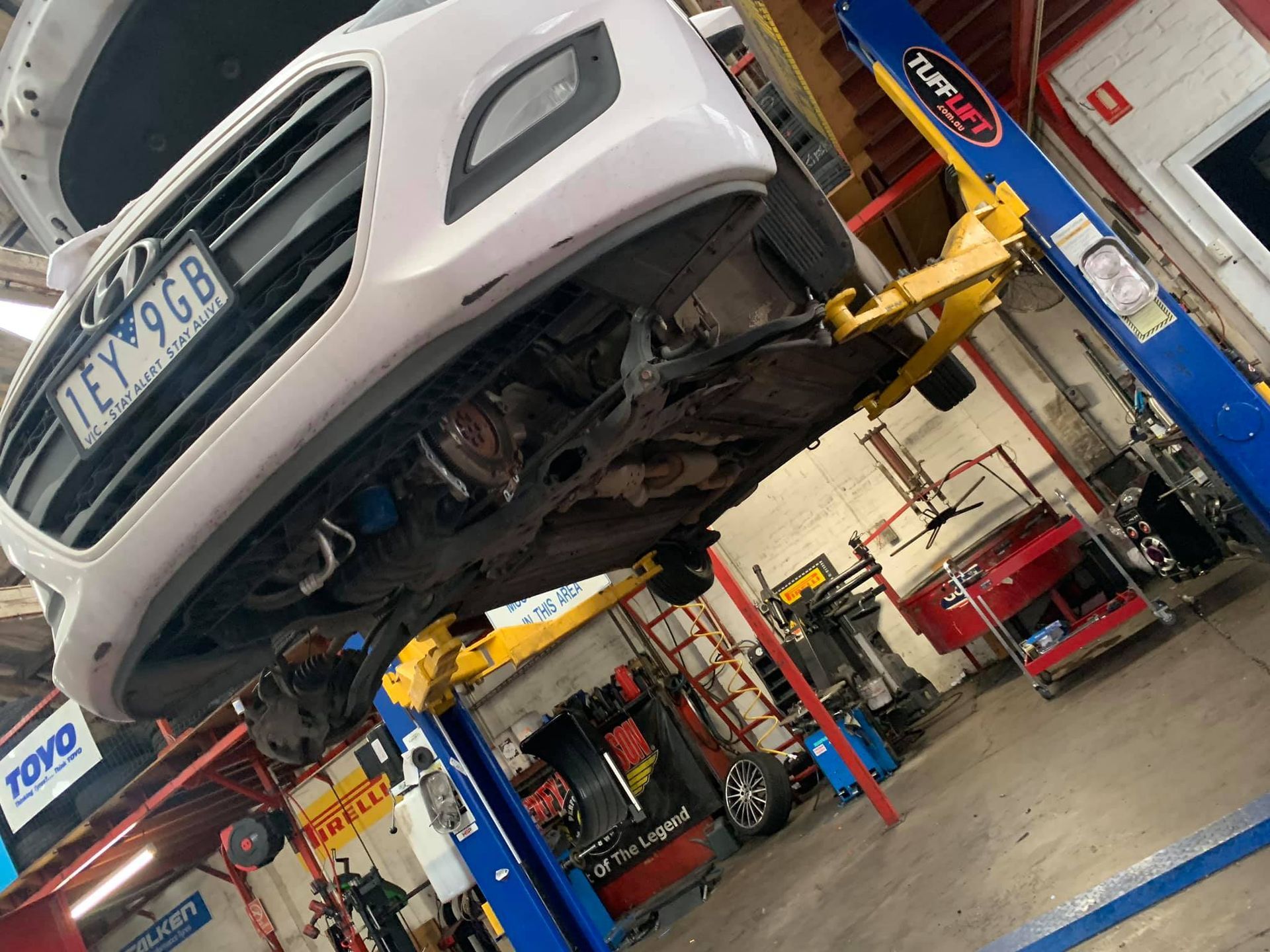 White car raised on a lift in a repair shop. Underside is visible; tools and equipment surround it.