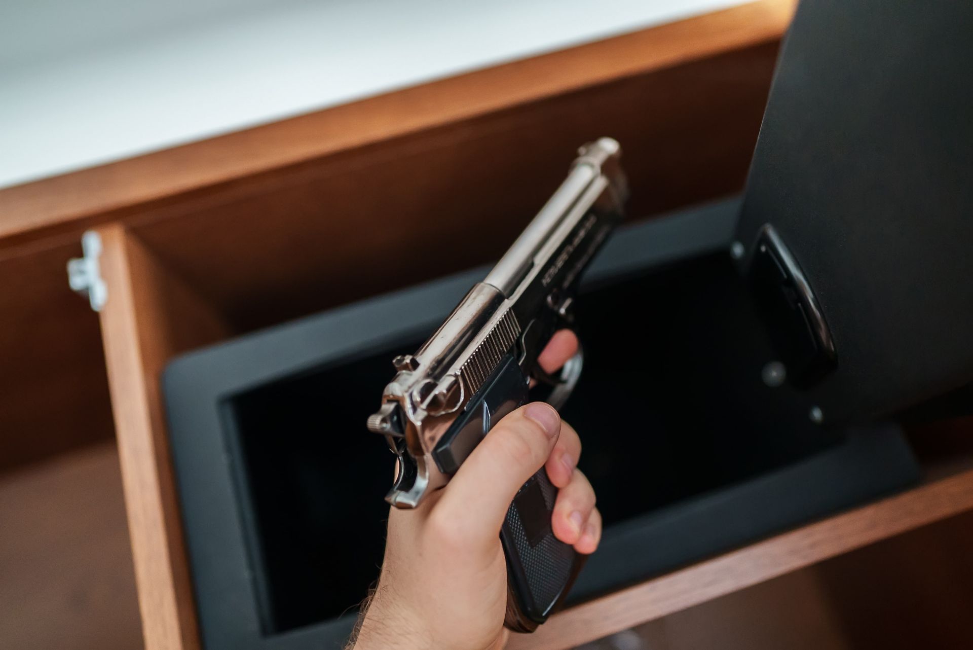 Hand holding a silver handgun, emerging from a black, open safe inside a wooden drawer.
