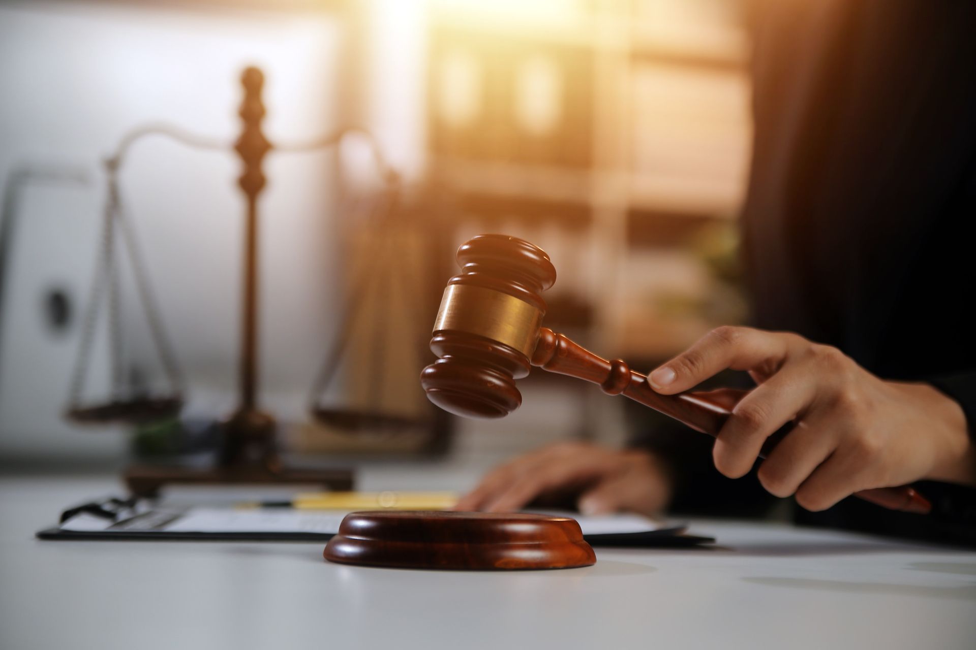 Person holding a gavel with scales of justice and documents on a white desk in a blurred office setting.