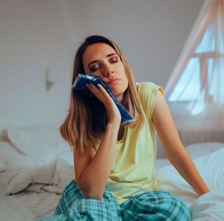 A woman is sitting on a bed holding an ice pack to her face