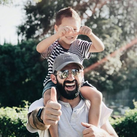 A man is carrying a little boy on his shoulders and giving a thumbs up.