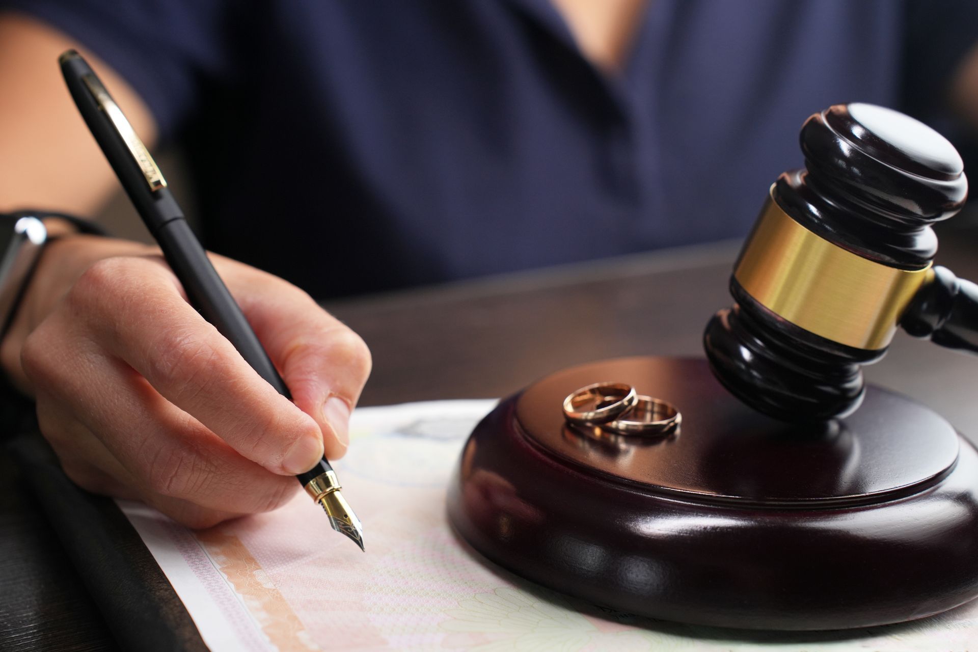 Hand writing with pen near gavel and wedding rings on a wooden base.