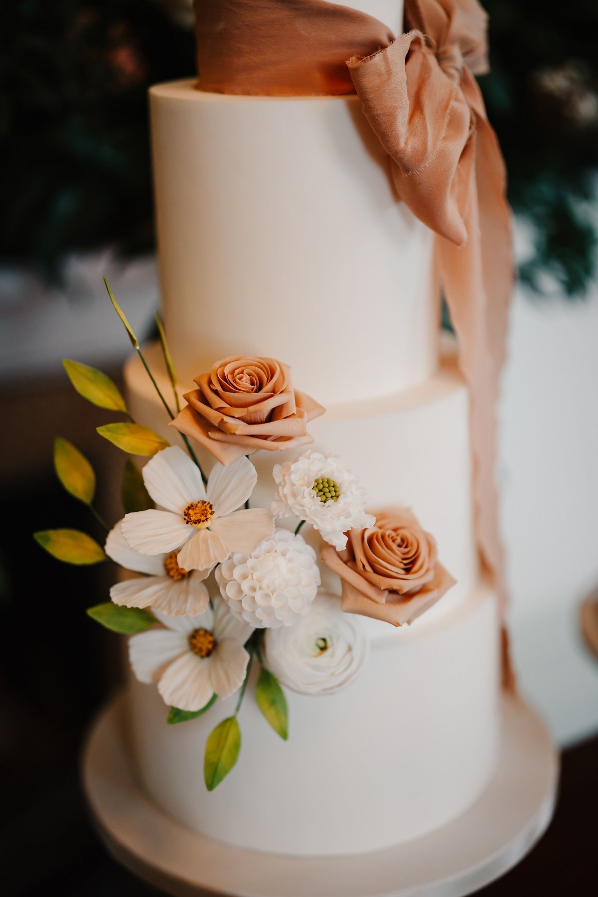 Three-tiered white cake with copper roses and other white and green floral decorations, brown ribbon, on a cake stand.