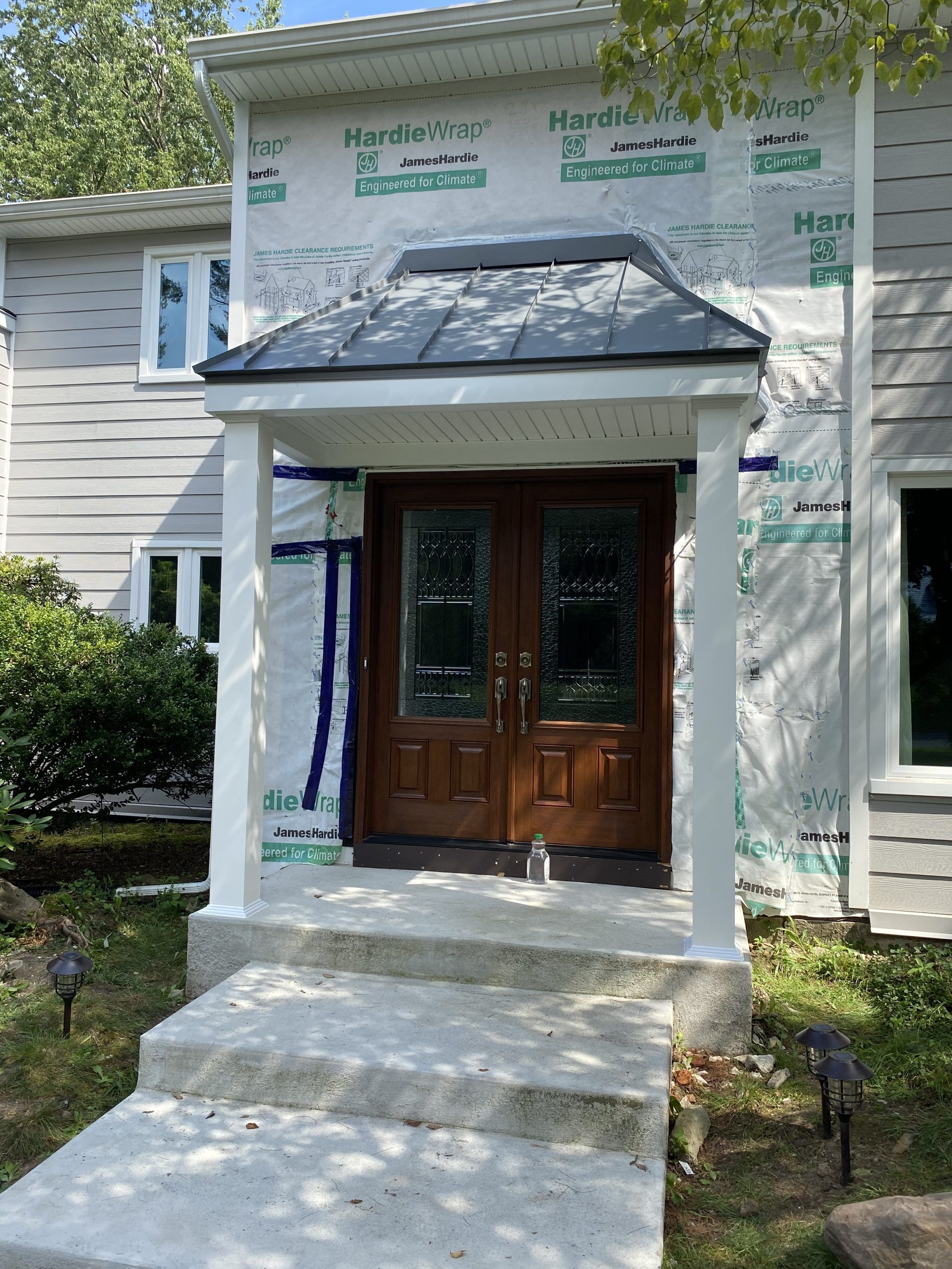 A two-story house with a wooden door under construction, covered with protective wrapping and white columns.