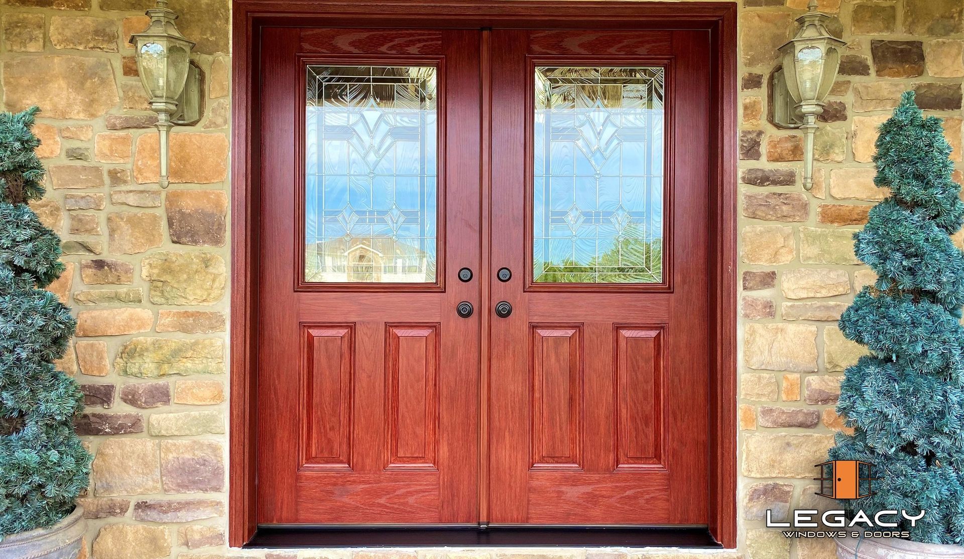 Red double doors with glass panels; flanked by topiaries and sconces, set in a stone facade.