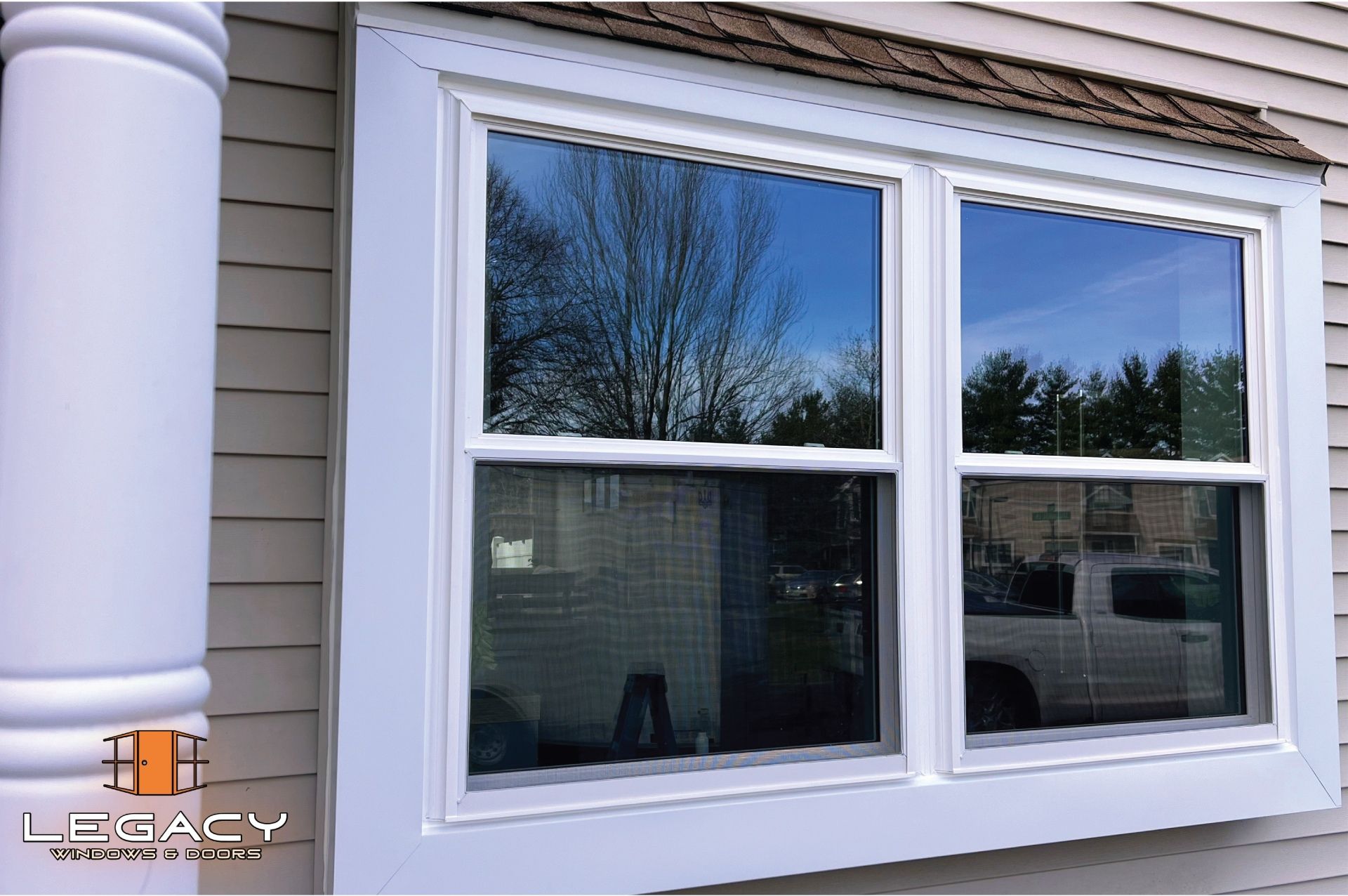 Double-hung windows in a white frame on a beige house, reflecting trees and a truck in the glass.