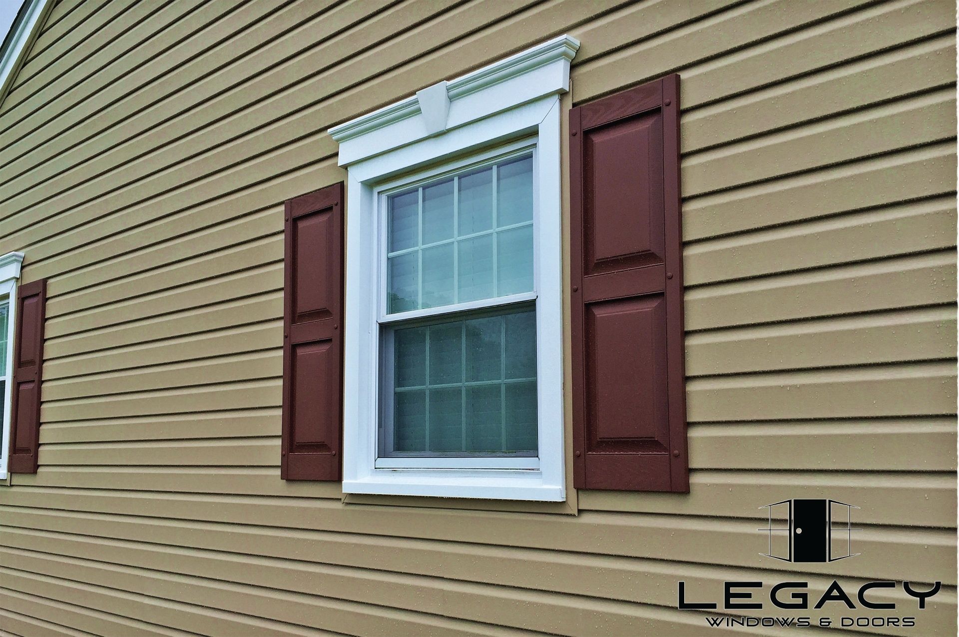 Tan siding with a window framed in white, burgundy shutters, and a decorative white header.