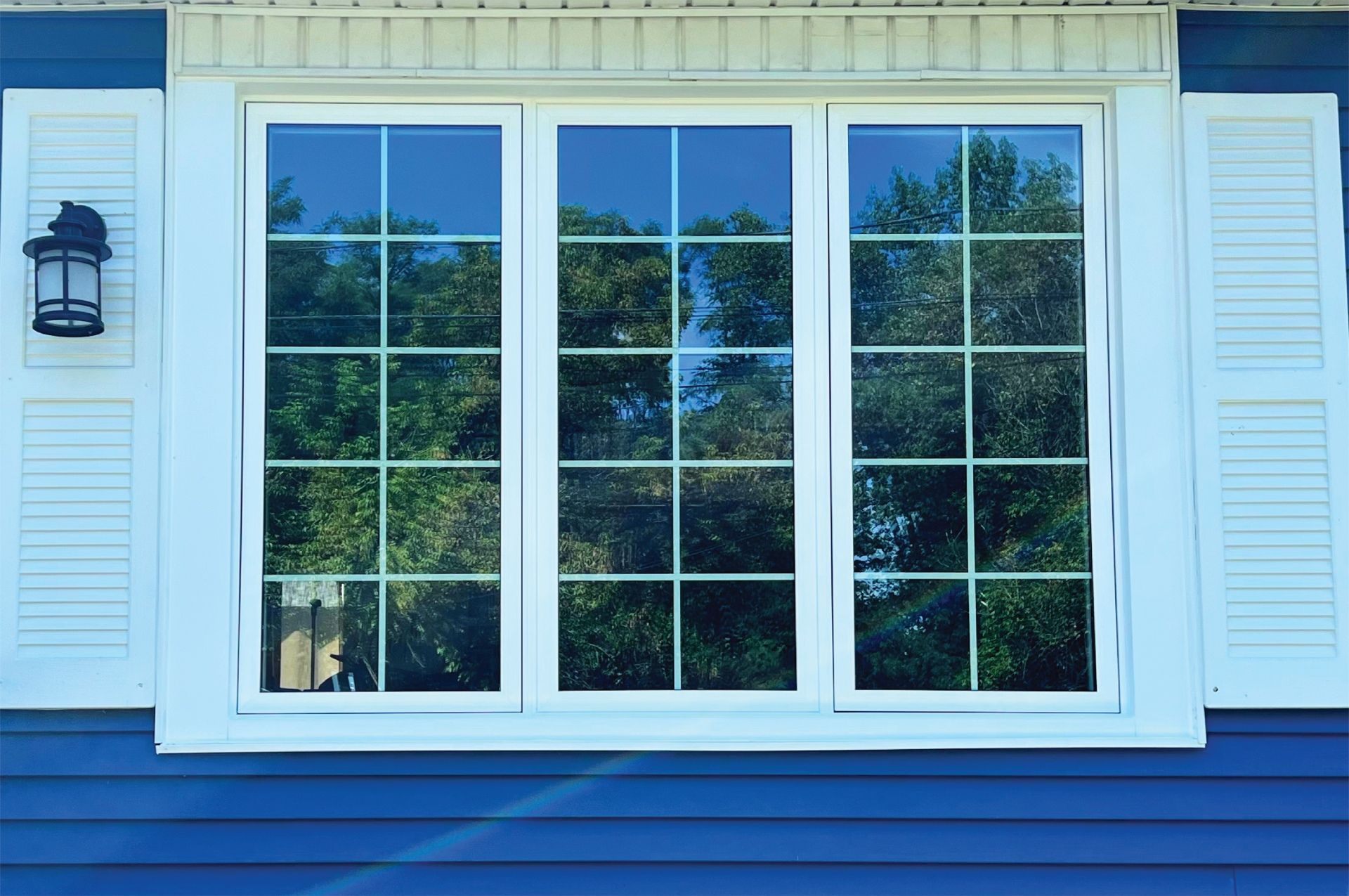 White-framed triple-pane window with grids reflects trees and sky, set against blue siding.