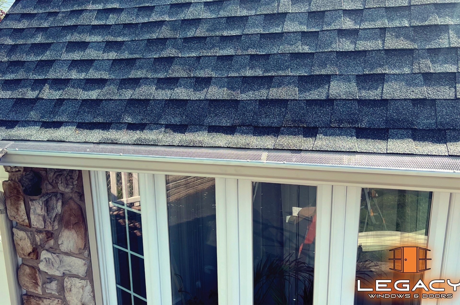 Close-up of a roof with gray shingles, gutters, and windows of a building with a stone exterior.