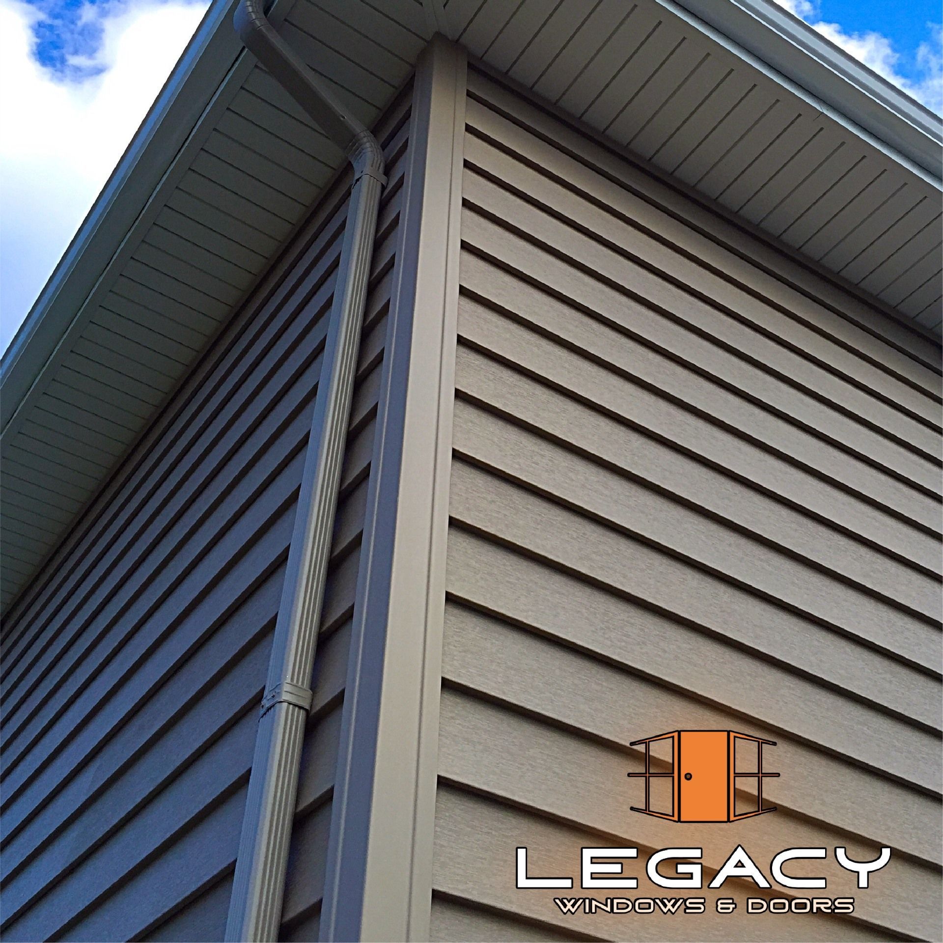 Corner of a house with gray siding, tan trim, and gutter against a blue sky.