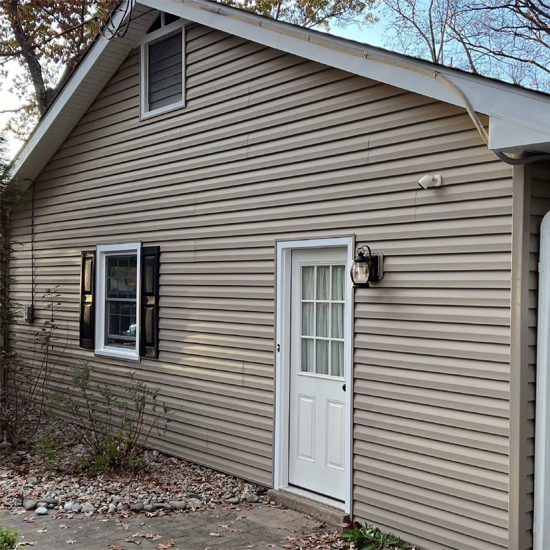 Tan-sided building with a white door, window with black shutters, and small window in the gable.