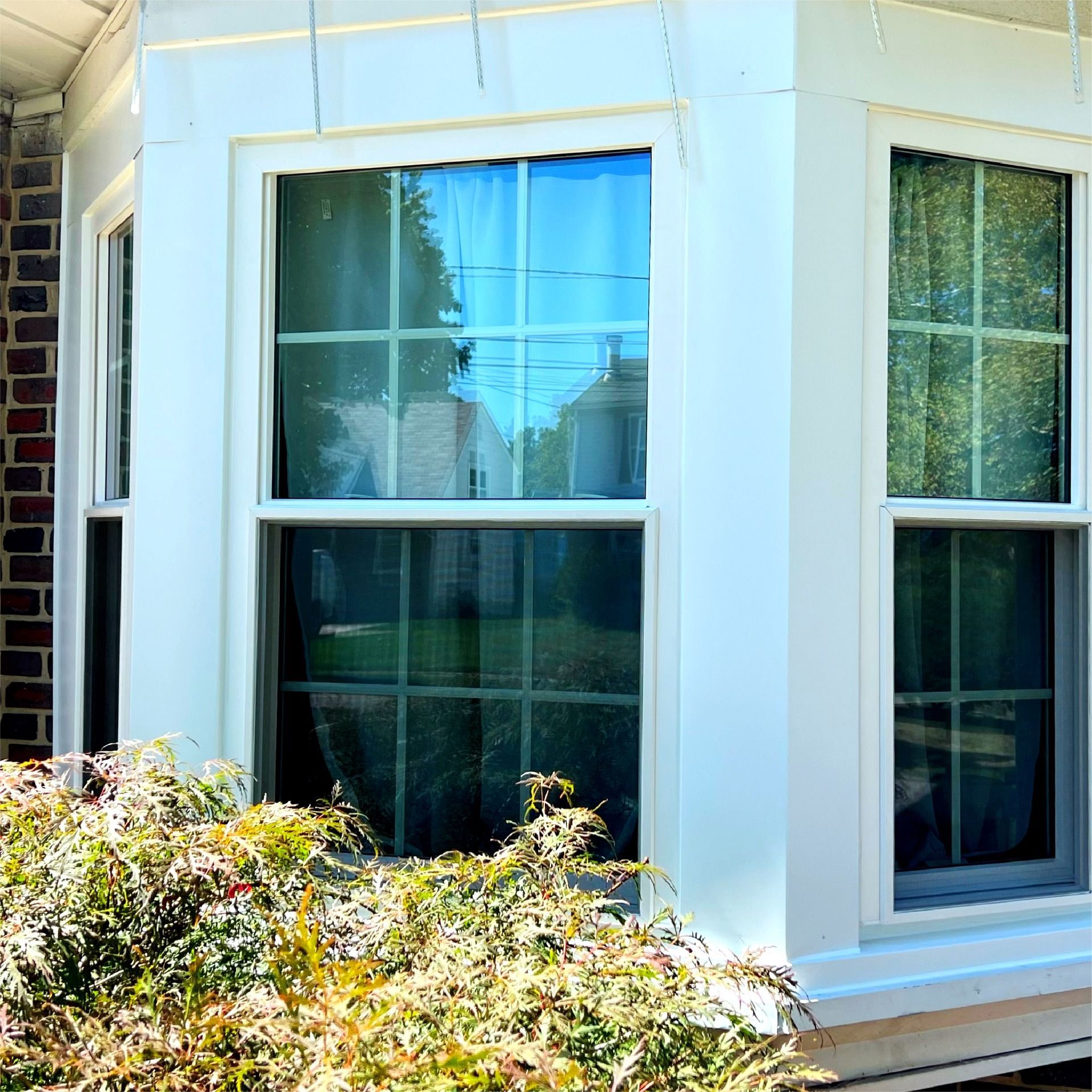 White bay window with clear glass panes on a brick building with bushes in front.