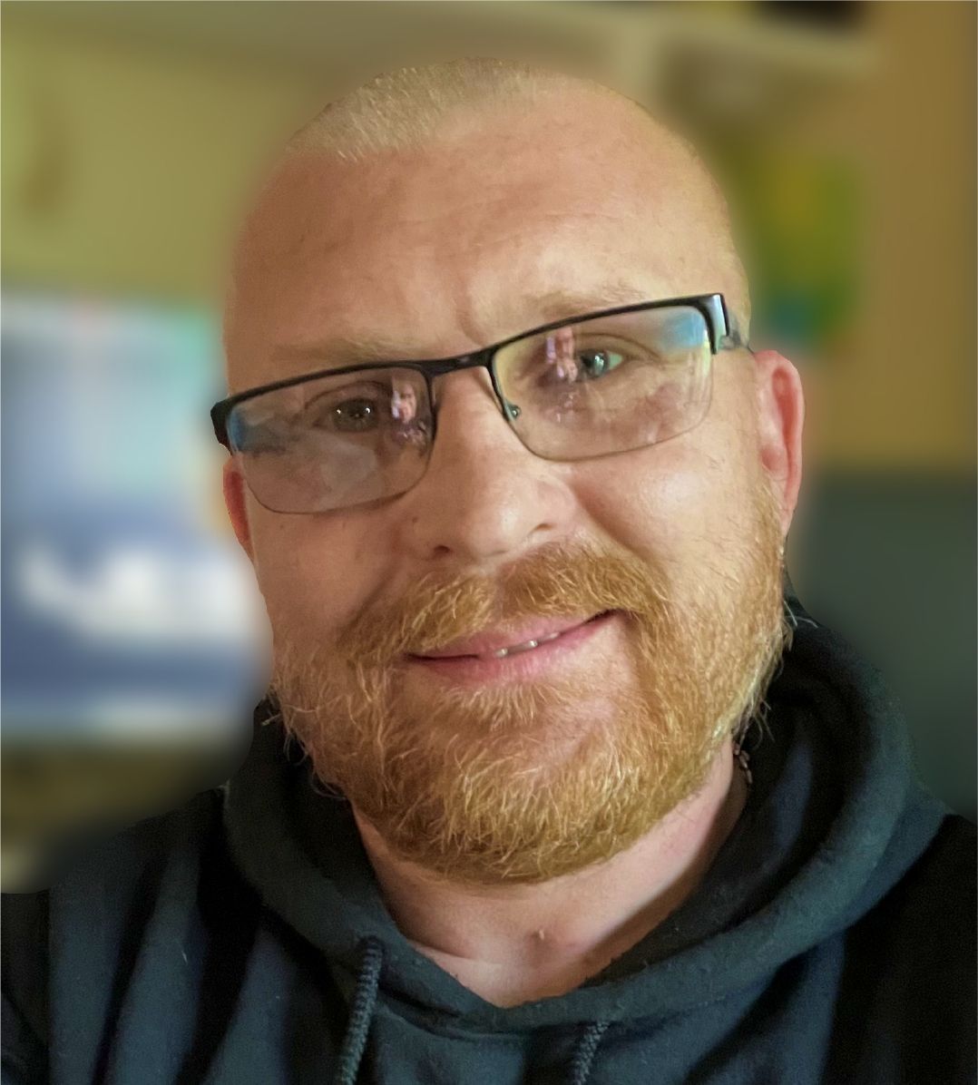 Man with a reddish beard and glasses, smiling, wearing a dark hoodie.