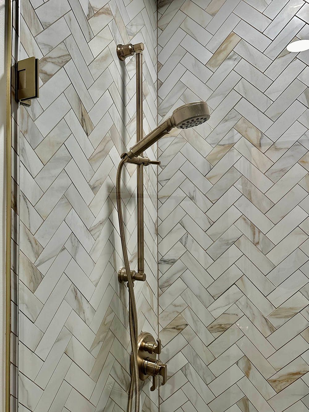 A shower with a herringbone tile wall and a brass shower head.