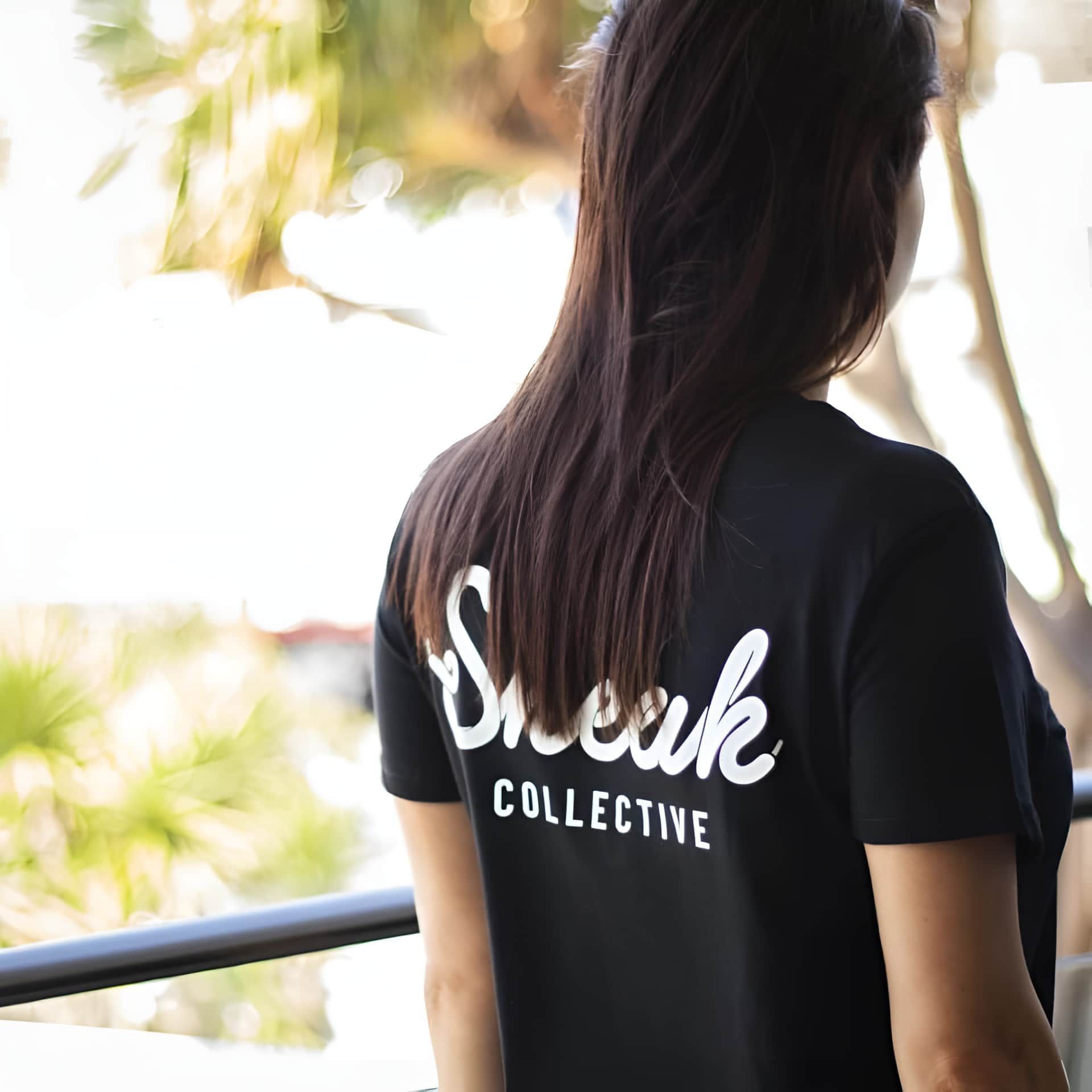 A Woman Wearing A Black Shirt That Says Sneak Collective — Carnival Clothing In Miami, QLD