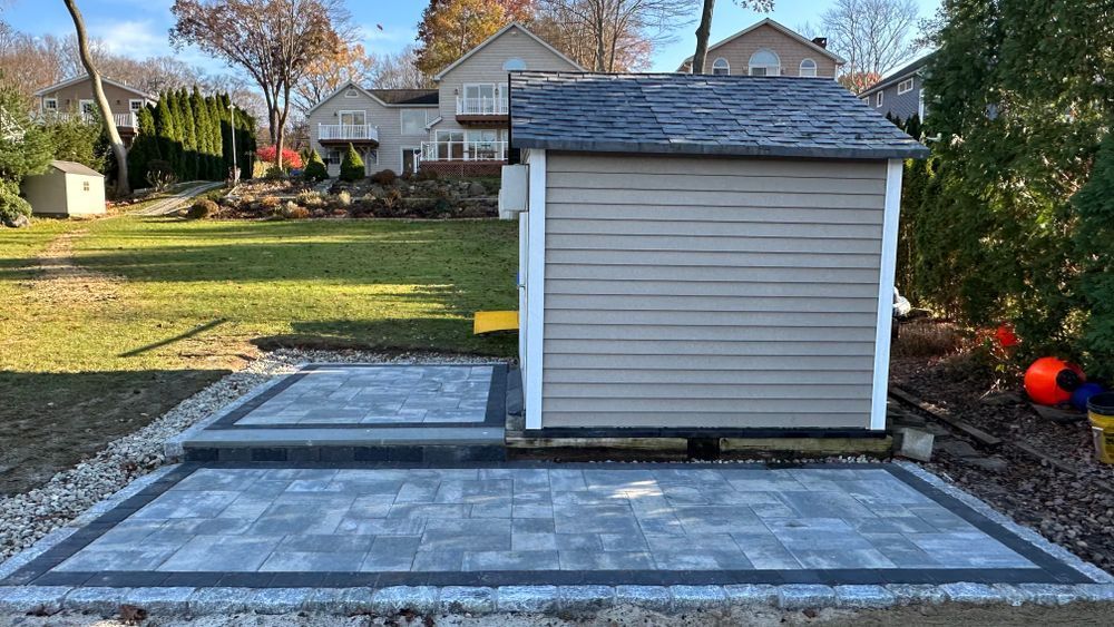 A backyard with a gray shed on a paver patio. A short step leads to the shed's base.