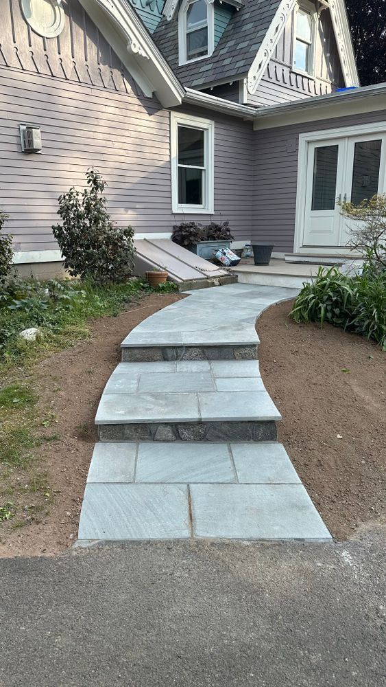 Stone steps leading to a house entrance with a grey facade and double white doors, surrounded by brown dirt and vegetation.