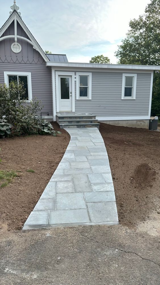 A stone pathway leads to a gray house with a white door and windows. The path is bordered by dirt and a lawn.