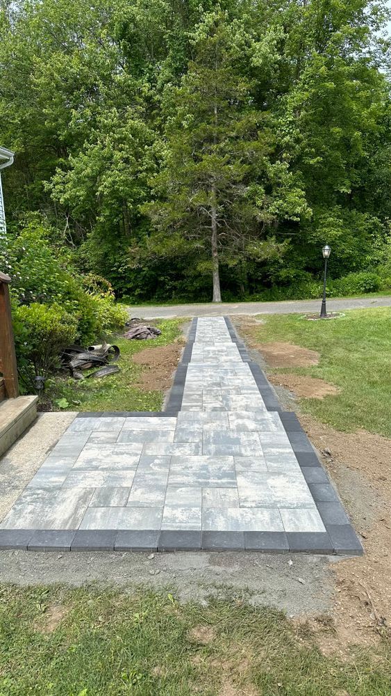 Stone paver walkway with dark border leading from a house entrance toward a road. Lush green trees and grass surround it.