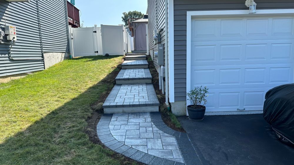 A paved walkway with steps leads uphill, next to a grassy slope and a garage. The pathway is lined with dark gray borders.
