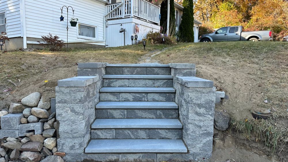 Stone steps leading up a hill towards a house, with a truck parked in the background on a sunny day.