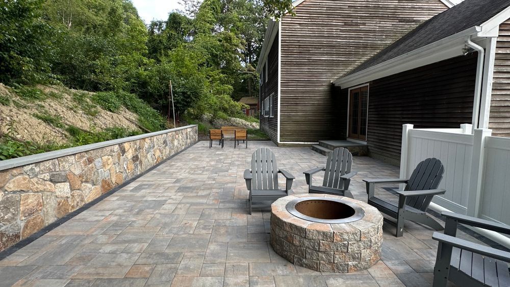 Patio with gray Adirondack chairs around a stone fire pit and a stone wall. A wooden house is in the background.