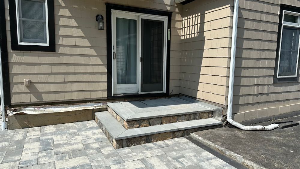Stone steps leading up to a sliding glass door on a beige shingled house. Paved walkway in front.