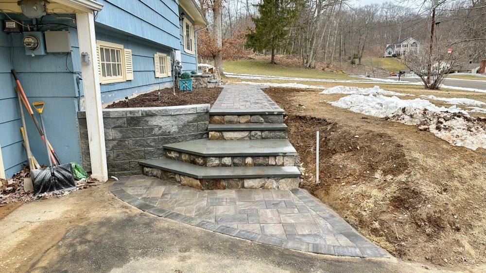 Stone steps and walkway leading up to a blue house with a partially snow-covered yard and trees in the background.