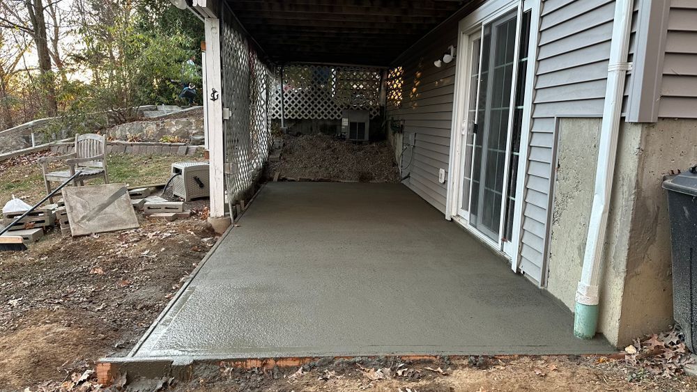 Newly poured concrete patio under a covered porch, next to a house with siding. The ground is bare around the patio's edges.