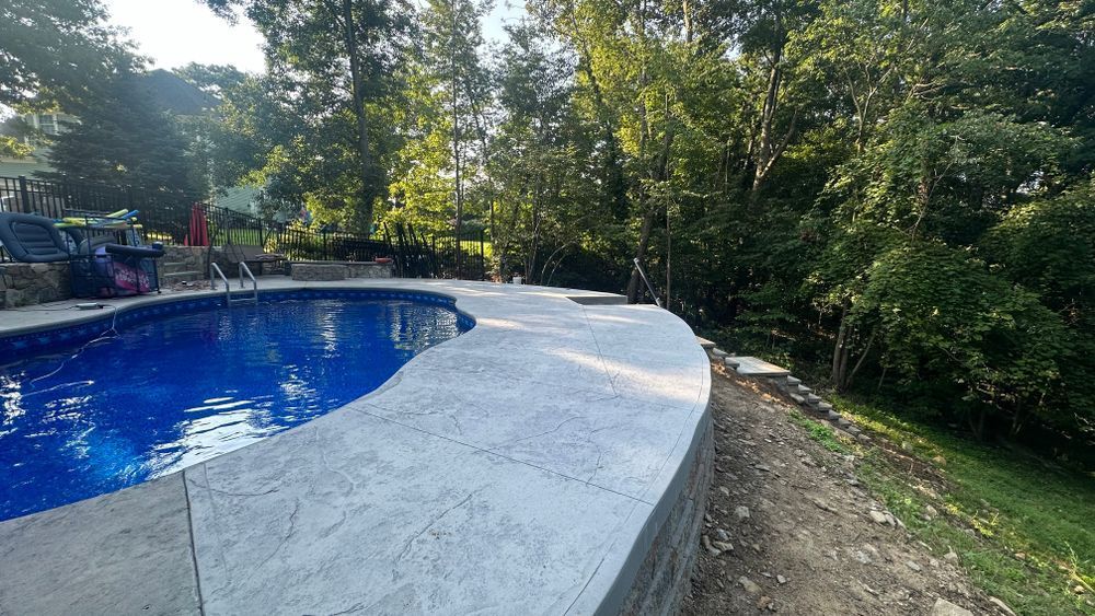 A backyard with a blue swimming pool and a gray concrete deck, bordered by greenery and trees on a sunny day.