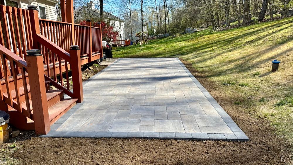 Paver patio extending from a wooden deck, surrounded by grass and dirt, against a hillside.