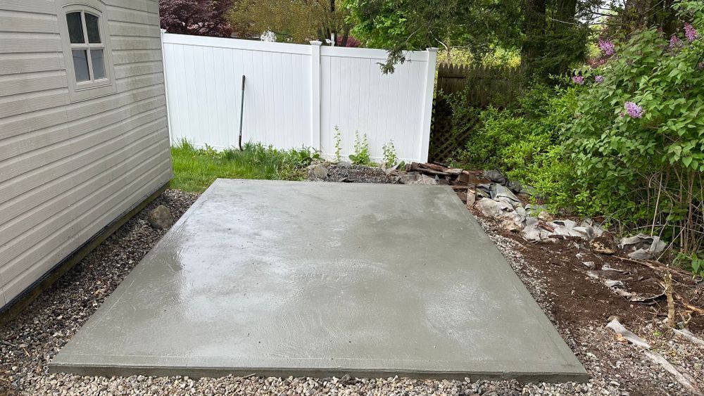 A newly poured concrete pad sits next to a white shed and a gravel base, with a white fence and greenery in the background.