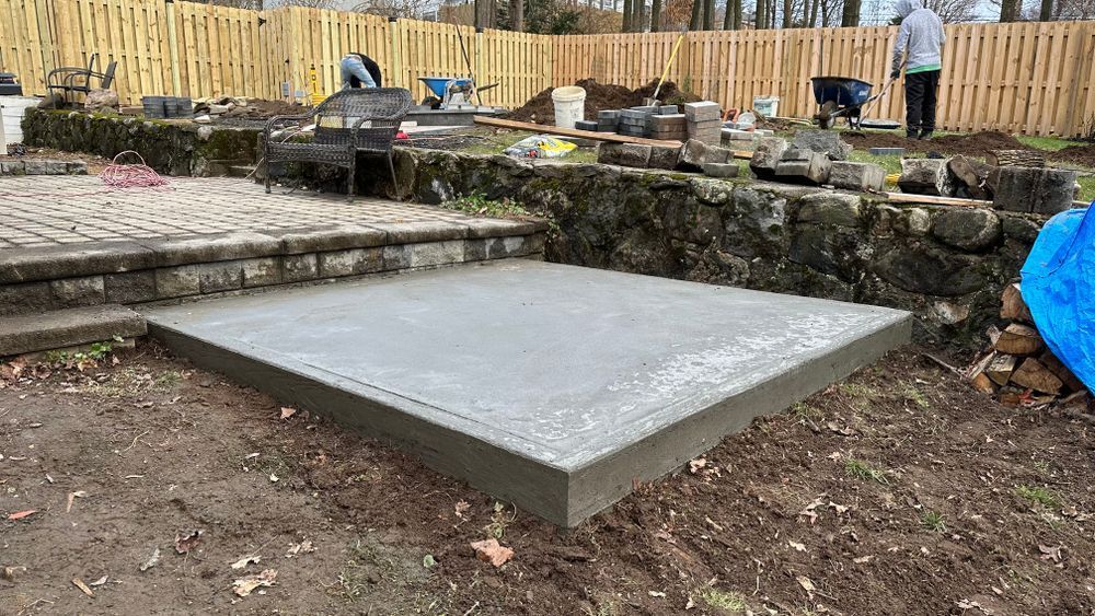 Concrete patio construction: freshly poured gray concrete slab in a backyard, with workers and building materials in the background.