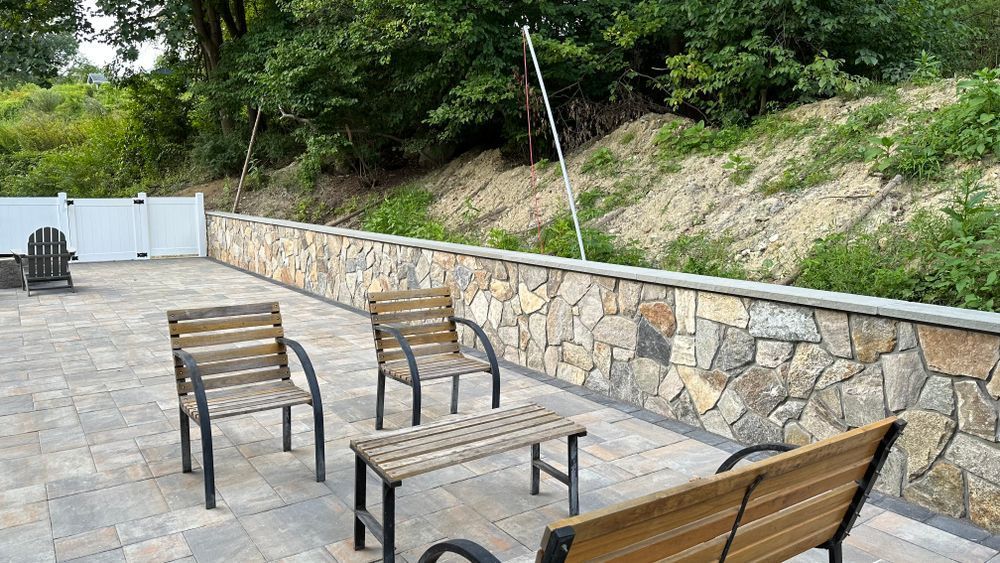 Patio with stone wall, three chairs, a bench, and a small table. The background has a hillside and white fence.