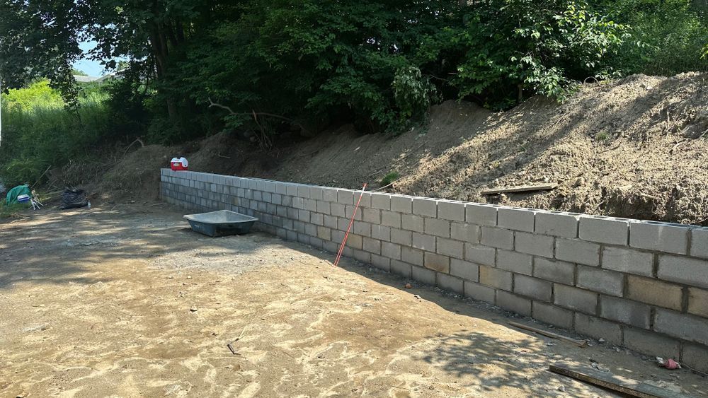 A gray cinder block retaining wall under construction on a dirt lot, backed by a hillside with greenery.