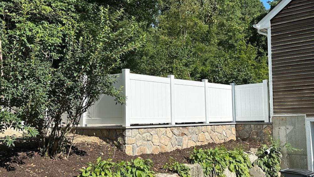 White vinyl fence atop a stone retaining wall in front of lush green trees and a house.