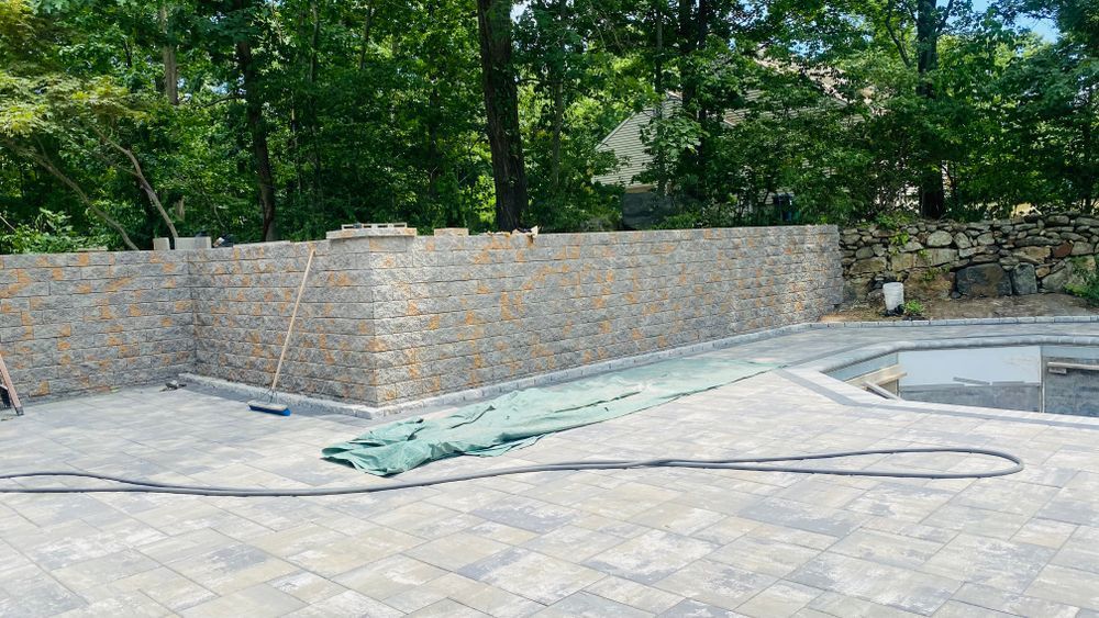 Stone wall beside a paved area with a green tarp on the ground, trees in the background.