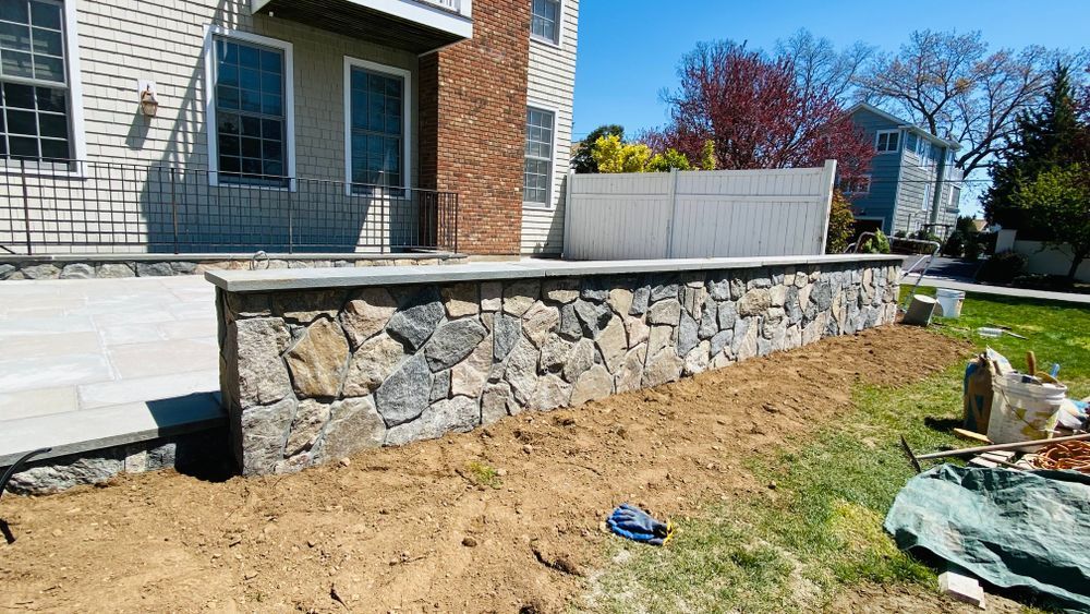 Stone retaining wall and patio bordering a house. Construction materials are visible in front of the wall on the grass, under a blue sky.