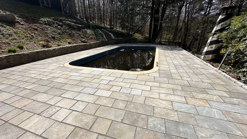 Stone-paved patio surrounds a dark, rectangular pool. The setting is outdoors, with trees in the background.