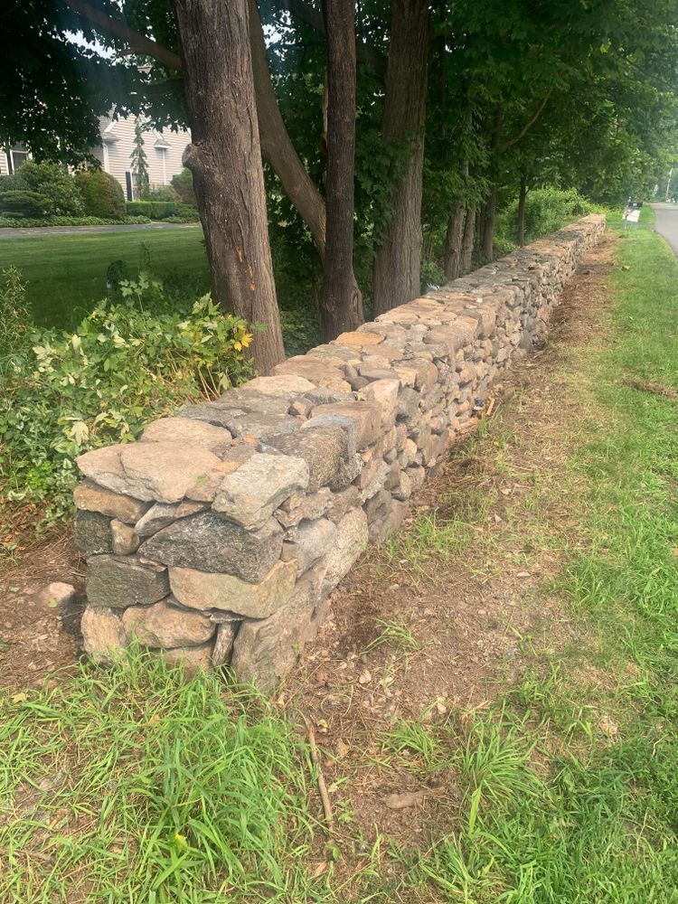 A low, dry-stone wall runs alongside a grassy area and road, with trees in the background.