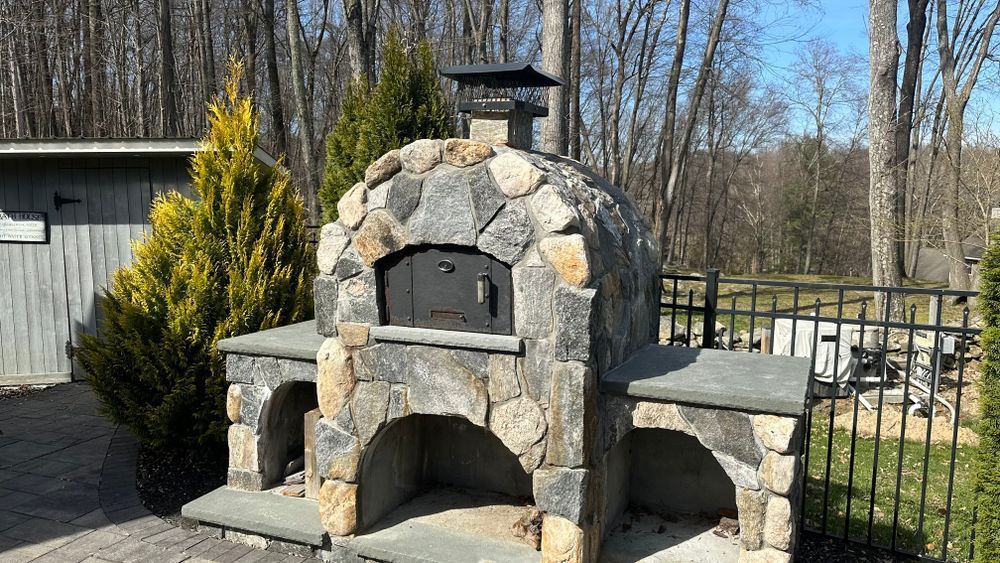 Stone pizza oven with arched openings, chimney, and surrounding greenery.